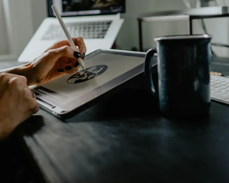 A person draws on a tablet with a stylus, surrounded by a laptop, keyboard, and mug on a dark desk, conveying focus and creativity in a digital workspace.