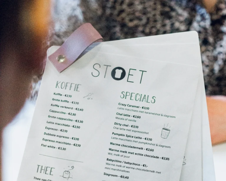 Close-up of a cafe menu titled "STOET," featuring sections for "Koffie" and "Specials." Prices are listed in Euros. A person sits in the background.