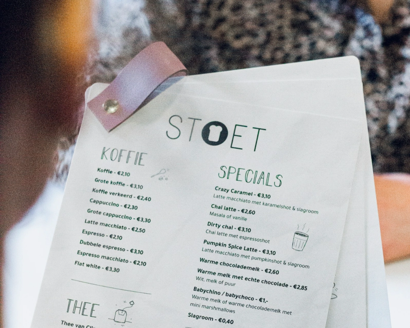 Close-up of a cafe menu titled "STOET," featuring sections for "Koffie" and "Specials." Prices are listed in Euros. A person sits in the background.