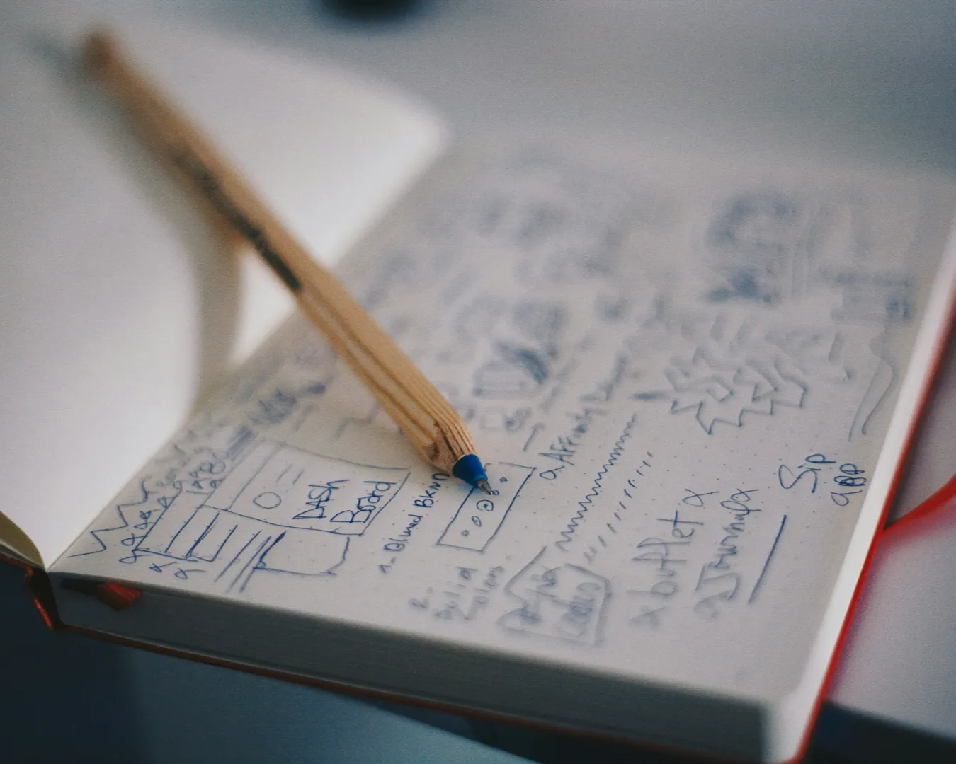 Open notebook with blue pen sketches and notes, a pencil resting on top. The scene conveys creativity and brainstorming in a casual setting.