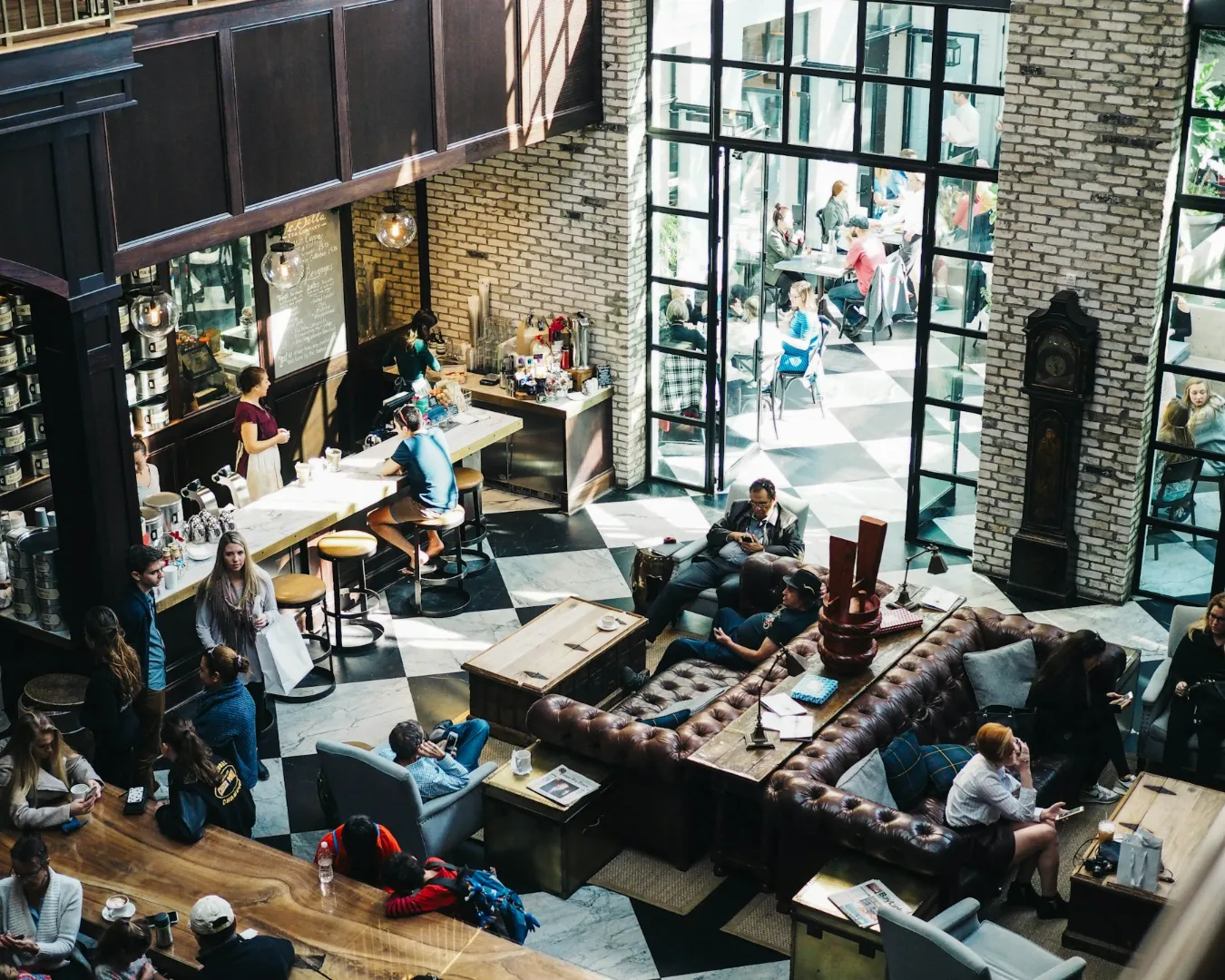 A bustling café interior with high ceilings and brick walls. Patrons relax on leather sofas and chairs, while others enjoy drinks at the bar. Sunlight streams through large windows, creating a warm, inviting atmosphere.