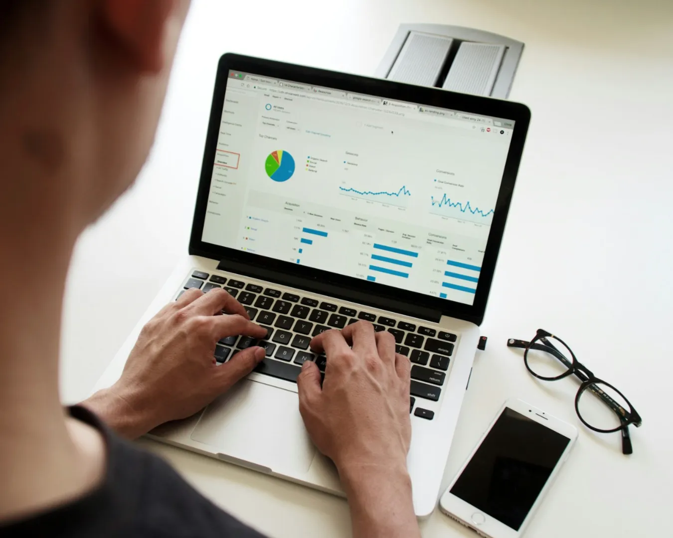 Person analyzing data on a laptop displaying charts and graphs, indicating business analytics. Nearby are glasses and a smartphone on a white desk.