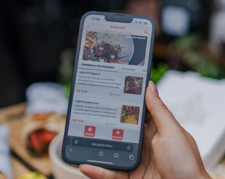 A close-up of a hand holding a smartphone that displays a food-delivery app, showing a Promos section, dish images, and Portuguese prices on the screen.