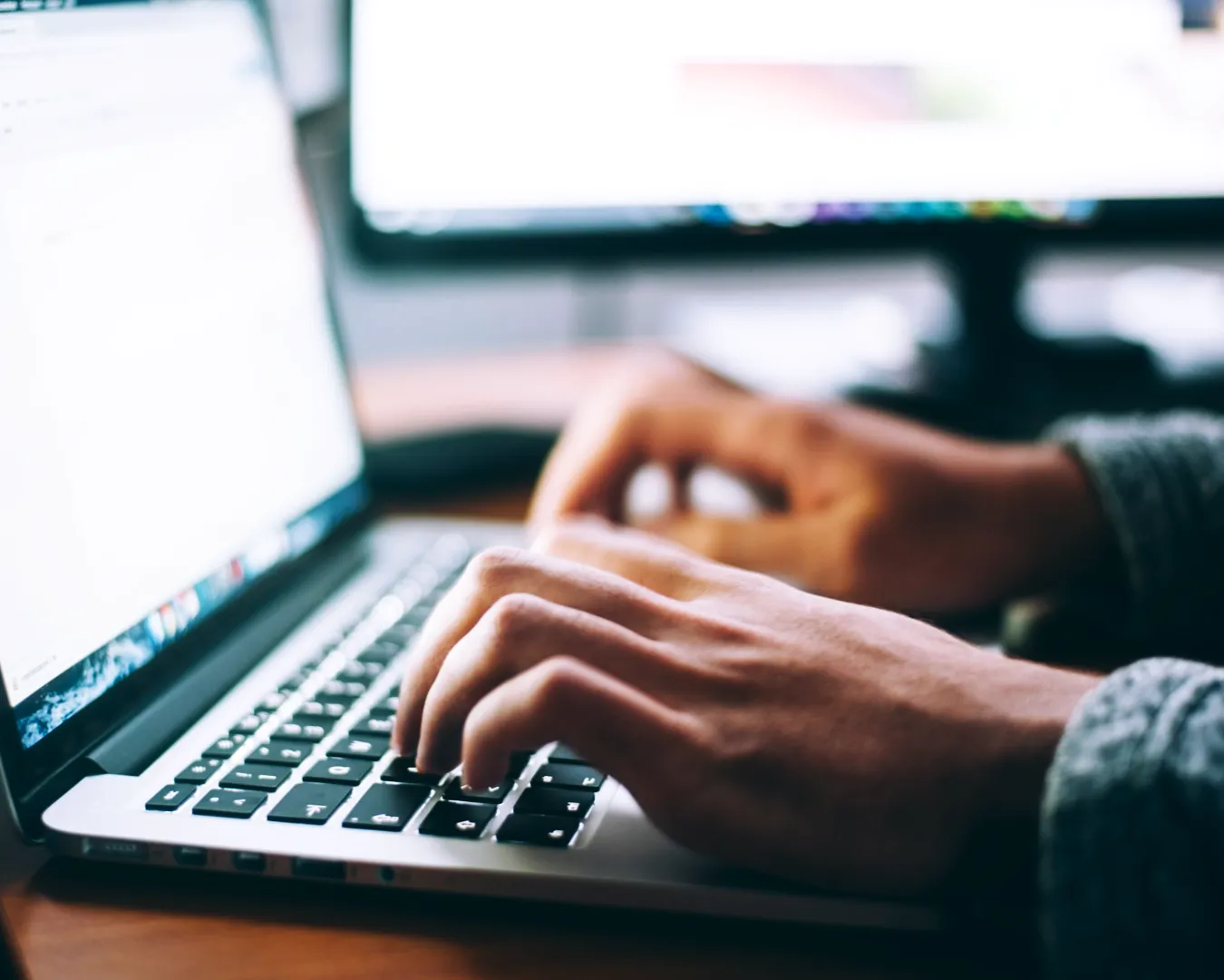 Hands typing on a laptop keyboard with a blurred monitor in the background. A focused, working atmosphere is conveyed.