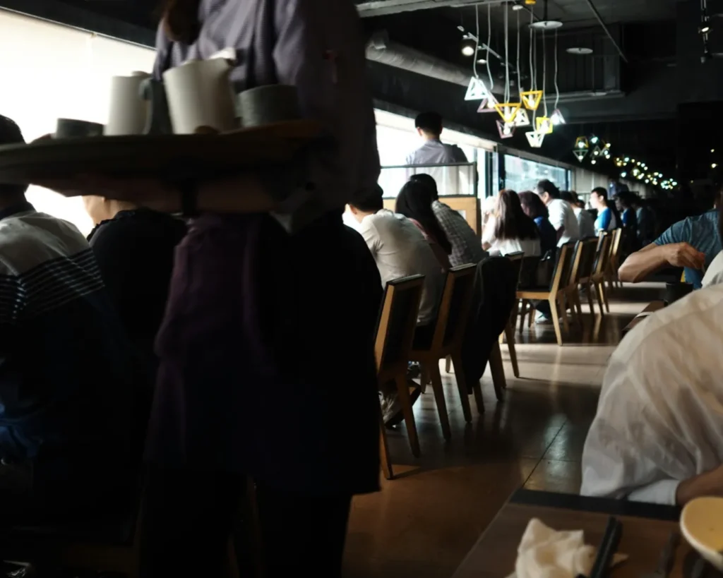 A busy cafe with people sitting at tables by large windows. A server carries a tray with cups. The atmosphere is lively with modern decor.