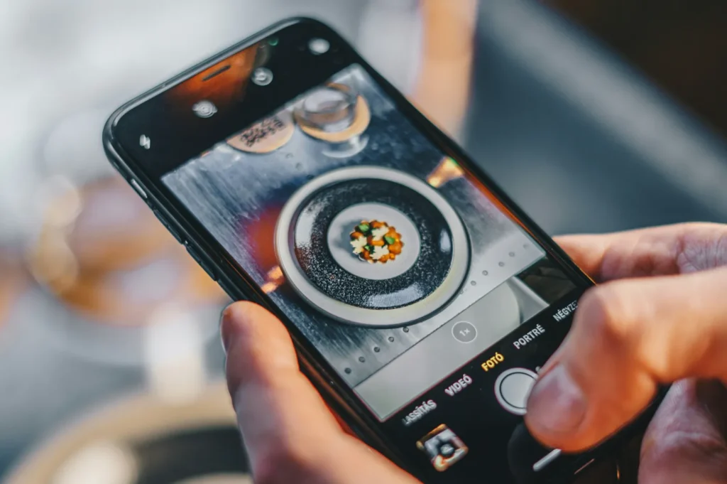 Close-up of hands holding a phone, capturing a gourmet dish with colorful garnishes on a plate. The scene is vibrant and artistic, highlighting food photography.