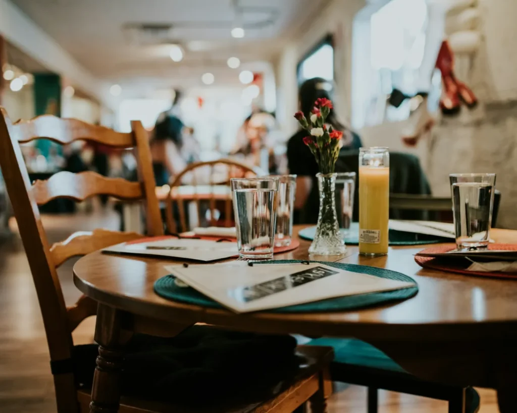A cozy restaurant setting with a round wooden table, empty glasses, a menu, a small vase with flowers, and a lit candle. The background shows blurred diners.
