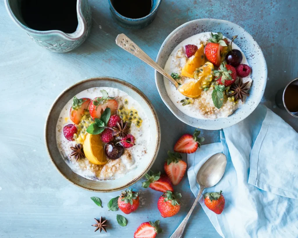 Two bowls of yogurt topped with vibrant fruit slices and star anise on a blue surface. Scattered strawberries and a cloth napkin add freshness.