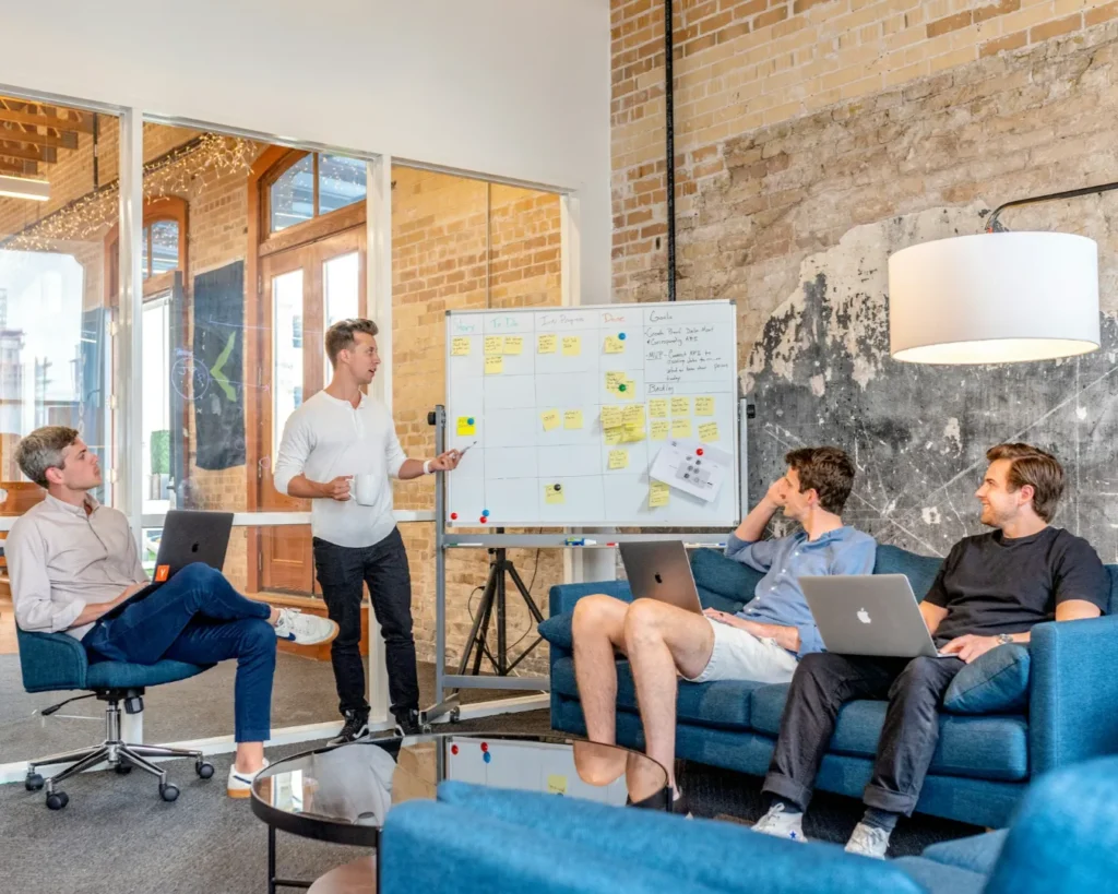 Four people in a casual meeting room engage in discussion. One stands by a whiteboard with sticky notes, while three others are seated with laptops.