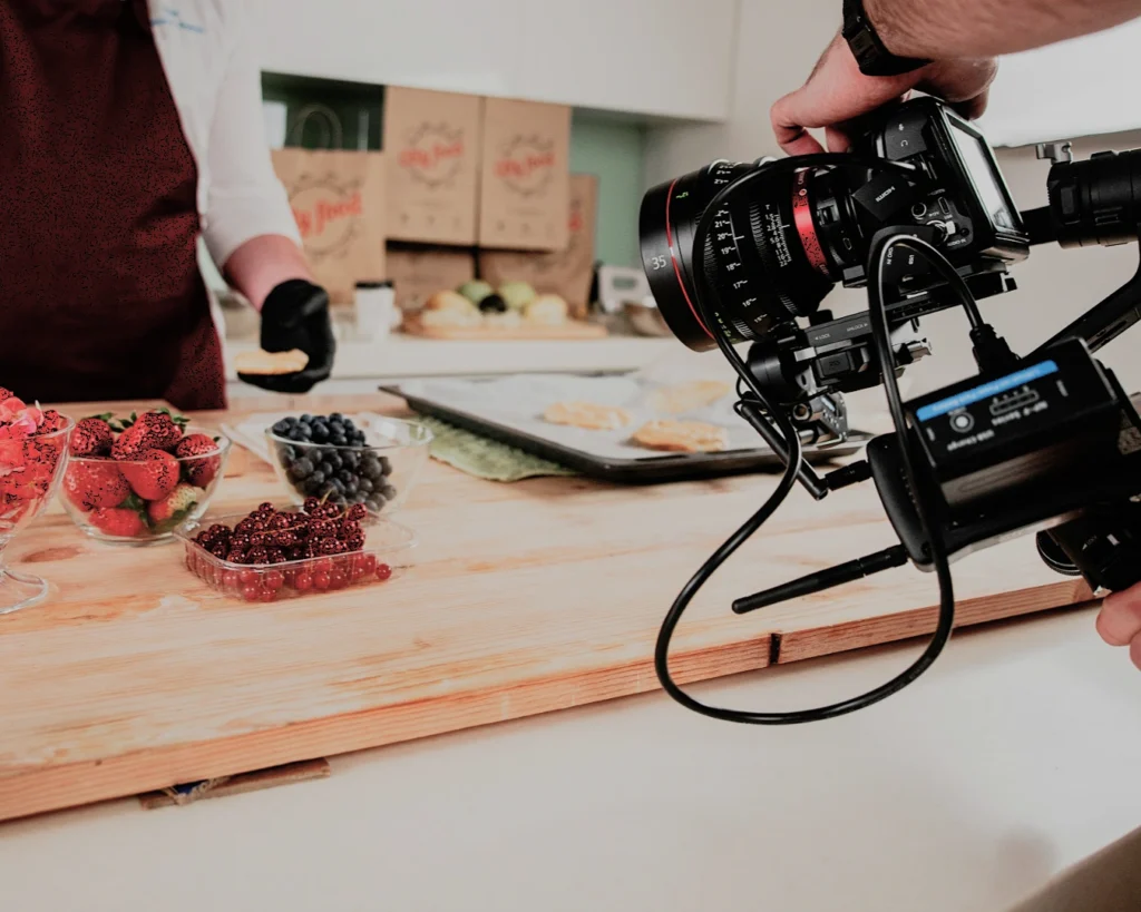 A person in a red apron is being filmed in a kitchen, preparing food. Fresh strawberries, blueberries, and other fruits are on the wooden table.