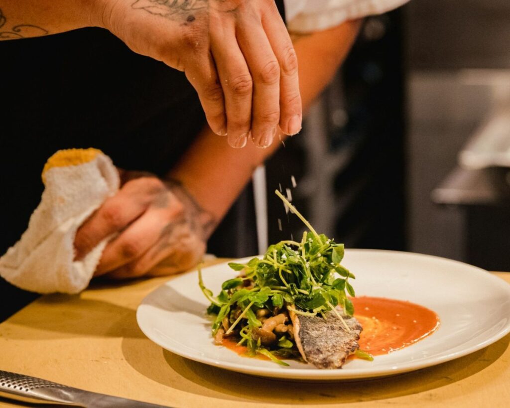 A chef's hand sprinkles seasoning over a plate of arugula-topped fish, resting on red sauce. The tattooed arm adds an artistic touch.