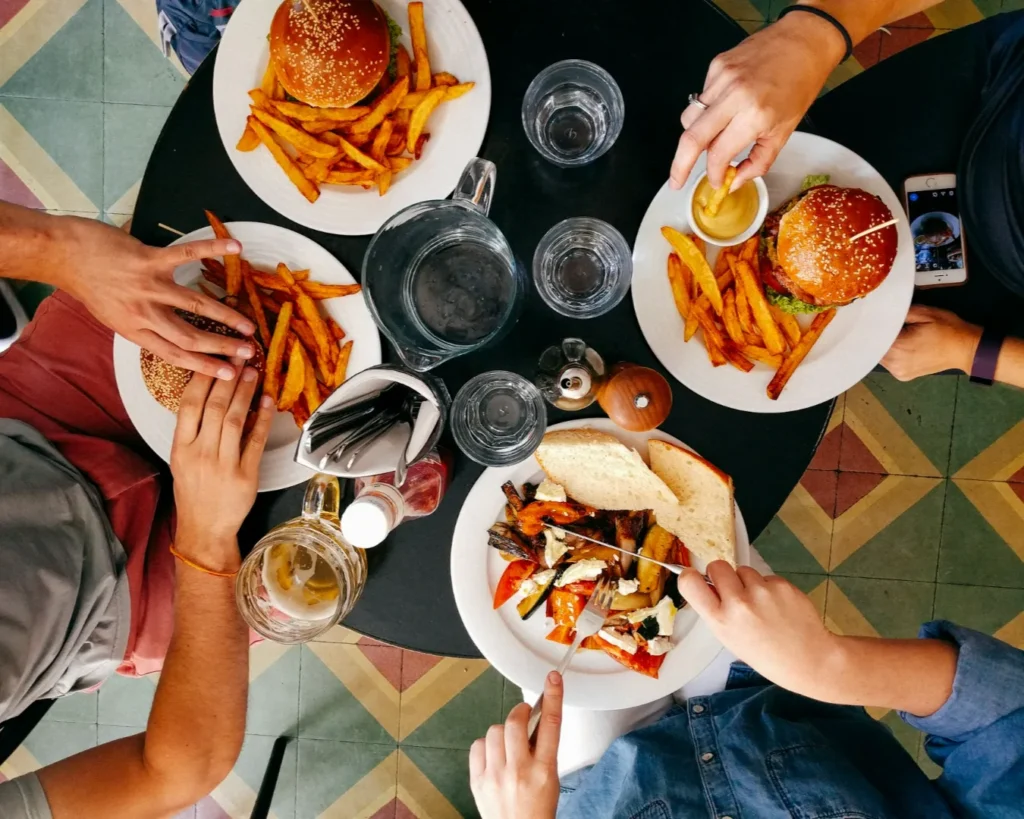 Aerial view of a round table with three people eating burgers and fries. One person uses a fork for grilled veggies. Casual and sociable dining scene.