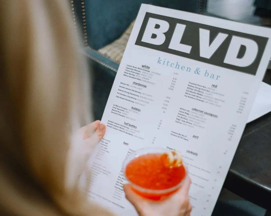 A person holds a cocktail and a menu titled "BLVD kitchen & bar" in a restaurant. The scene conveys a relaxed, social dining atmosphere.