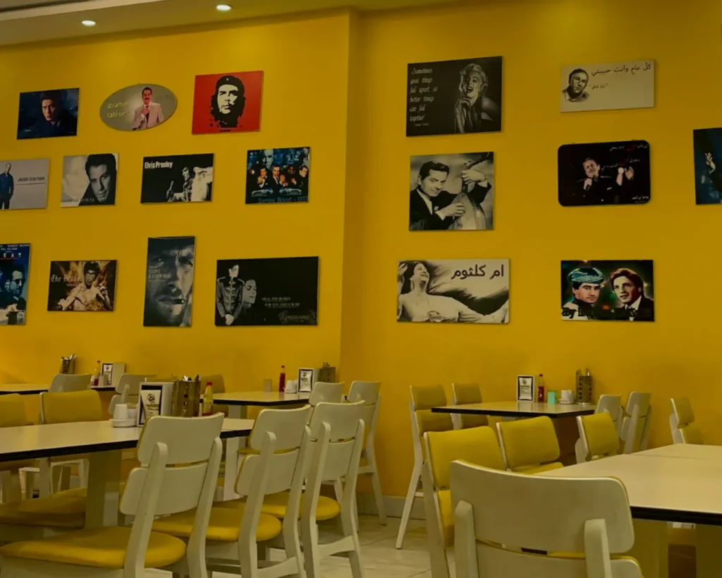 A cozy cafe with yellow walls adorned with vintage celebrity posters. White tables and chairs fill the space, creating a retro, welcoming atmosphere.