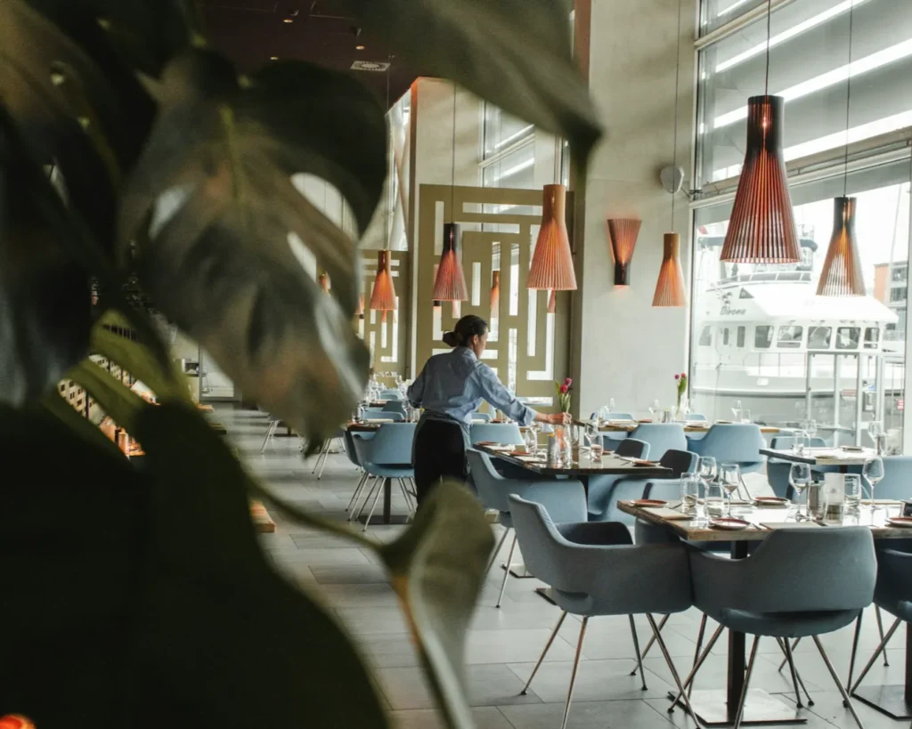 Elegant restaurant interior with modern decor, soft lighting, and large windows. A person sets tables with ocean views in the background, creating a serene atmosphere.