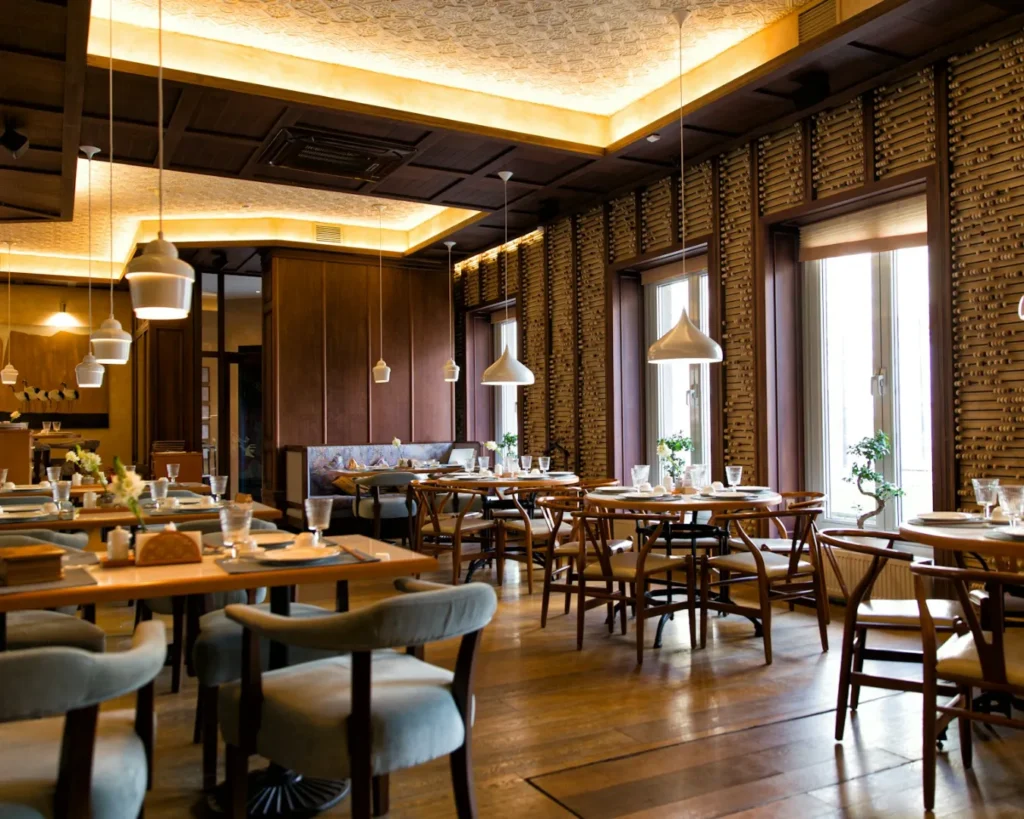 Elegant restaurant interior with wooden walls and floor, round tables, cushioned chairs, hanging lights, and large windows. Cozy and inviting atmosphere.