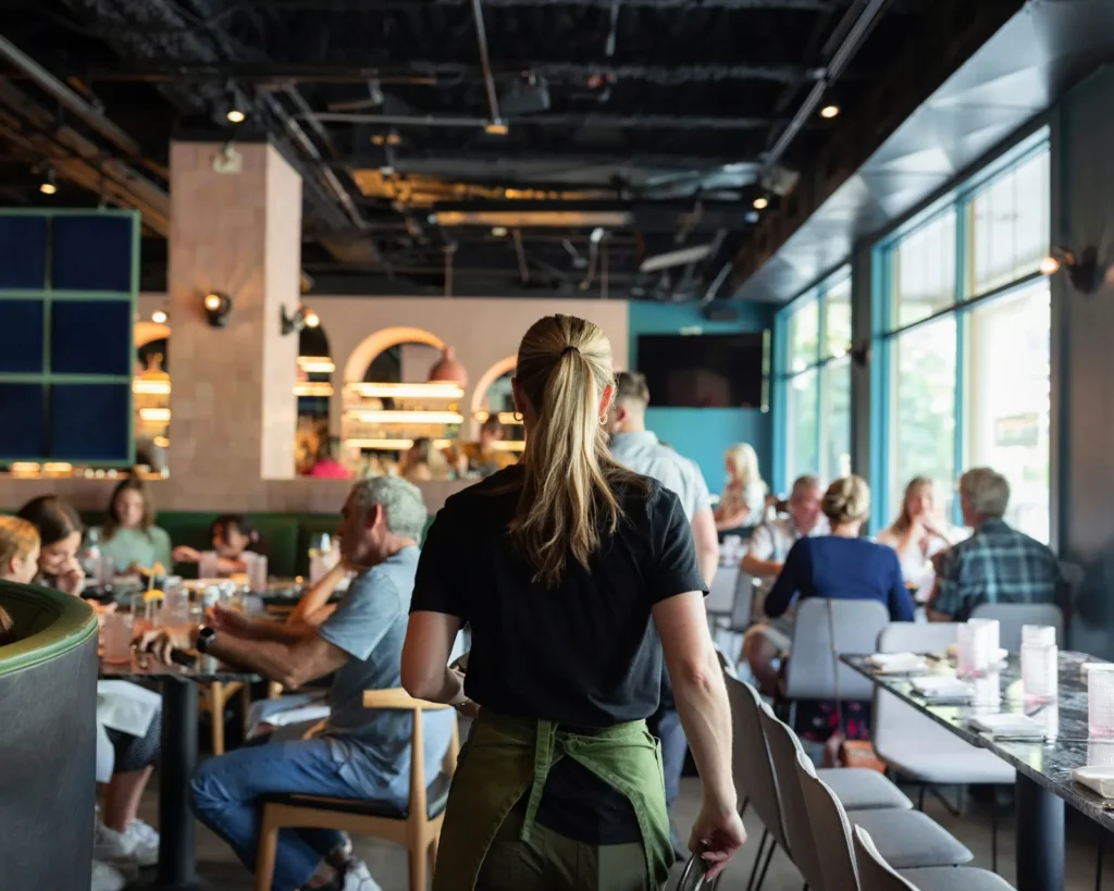 A waitress in a black shirt and green apron walks through a bustling restaurant. Diners sit at tables enjoying meals, with a lively and casual atmosphere.