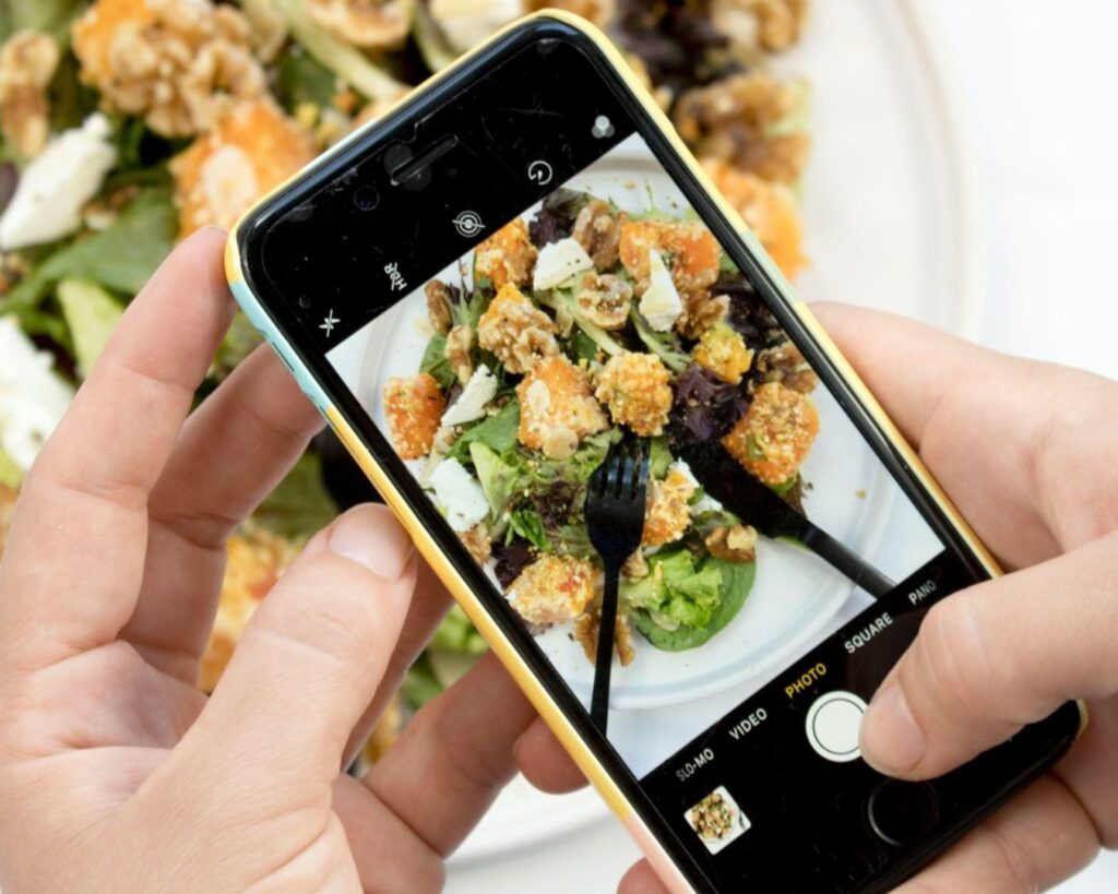 A person holds a smartphone above a plate of salad, capturing a close-up shot of greens, croutons, feta, and walnuts with the device's camera.
