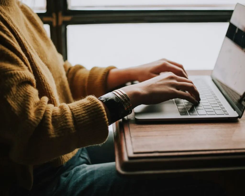 A person in a cozy yellow sweater types on a laptop at a wooden desk. Sunlight filters through a window, creating a warm and focused atmosphere.
