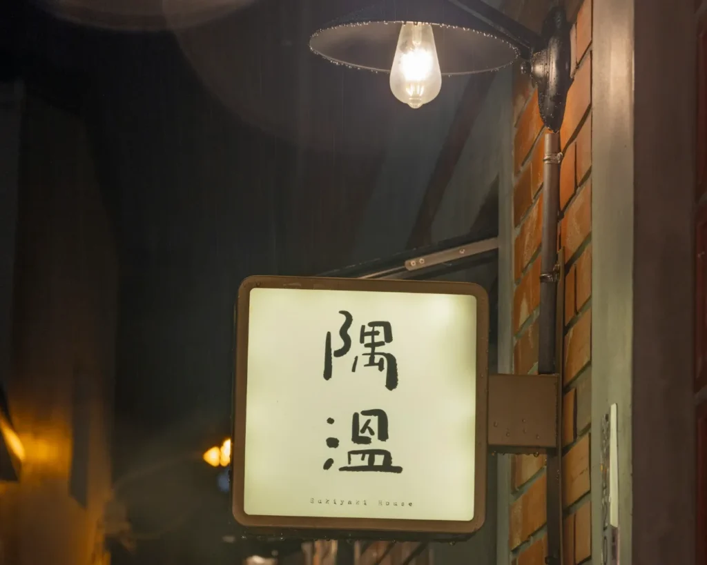 Lit at night, a rectangular sign with black Japanese characters and "Sukiyaki House" in English hangs on a brick wall under a glowing bulb.