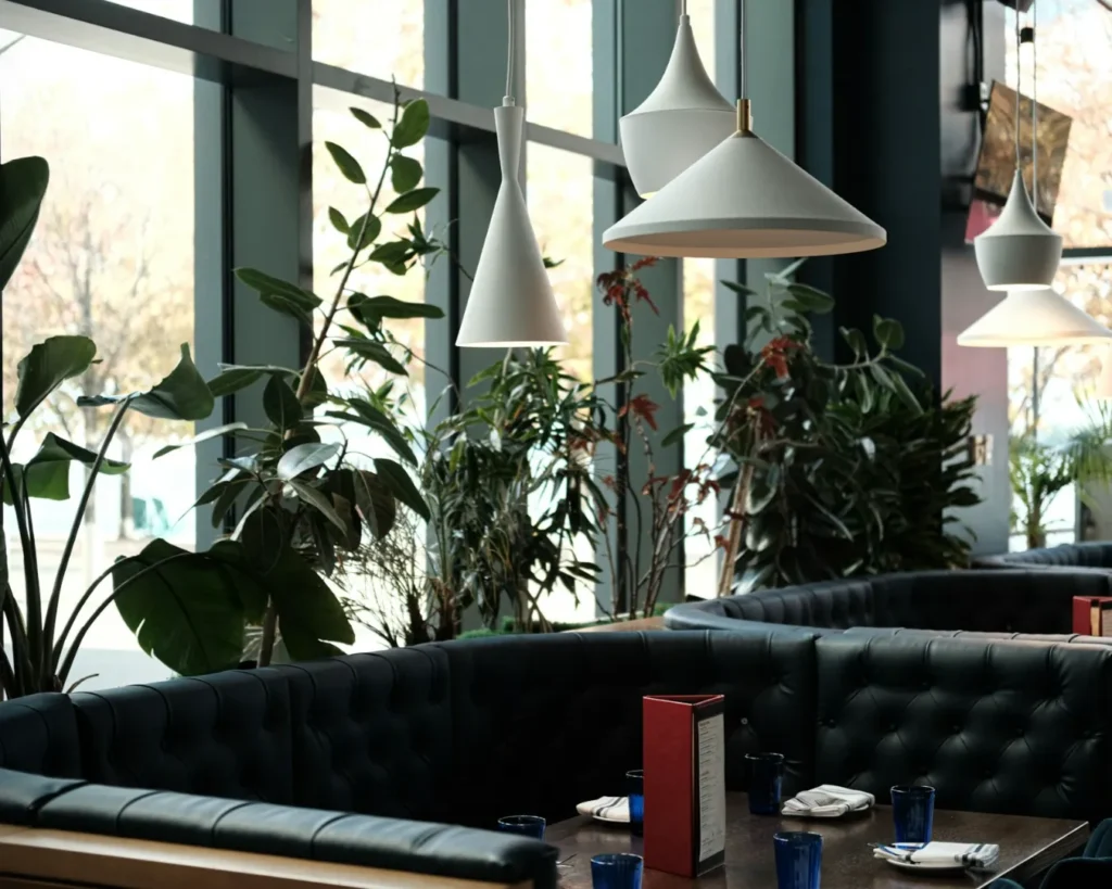 Modern restaurant interior with dark teal, tufted booth seating and white pendant lights. Green plants line large windows, creating a cozy atmosphere.