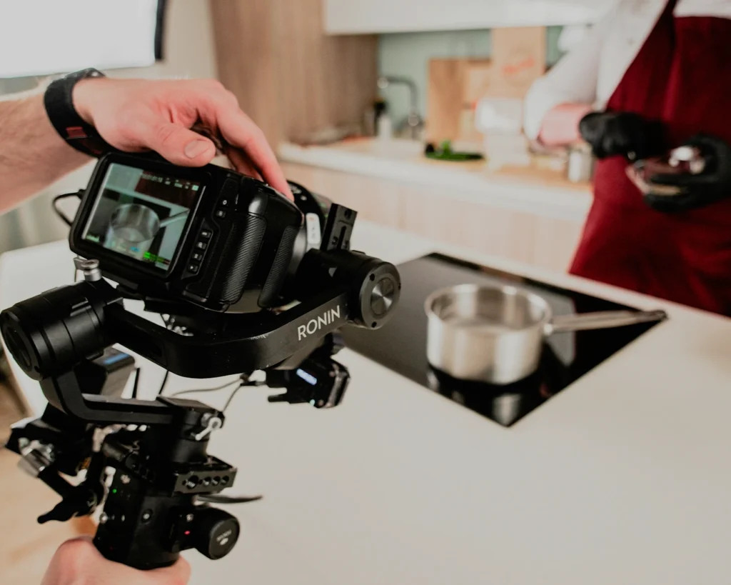 A person operates a camera rig in a modern kitchen to film a cook wearing a red apron. A pot simmers on an induction stove.