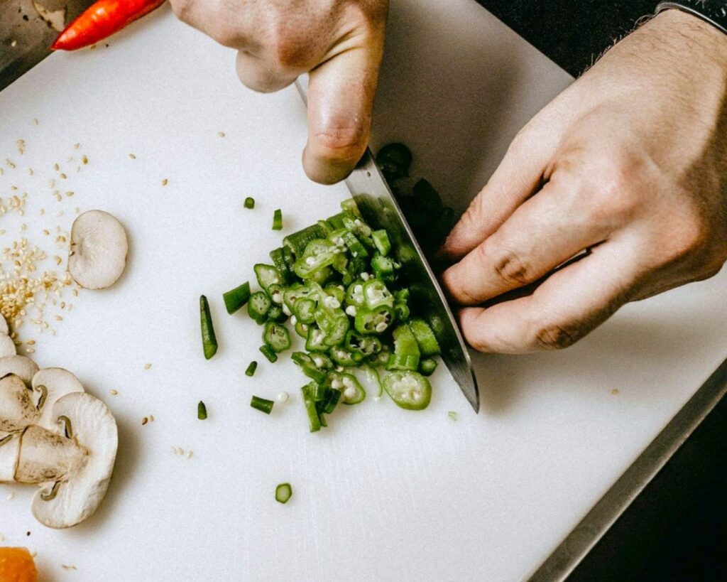 A person is chopping green okra on a white cutting board. Nearby, sliced mushrooms, sesame seeds, and a red chili pepper are arranged. The tone is focused and culinary.