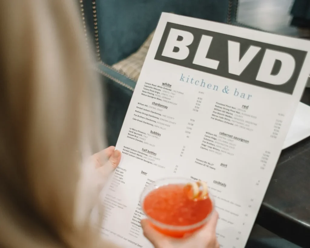 A person holds a menu from "BLVD kitchen & bar" in one hand and a red cocktail in the other. The setting is a cozy restaurant with muted lighting