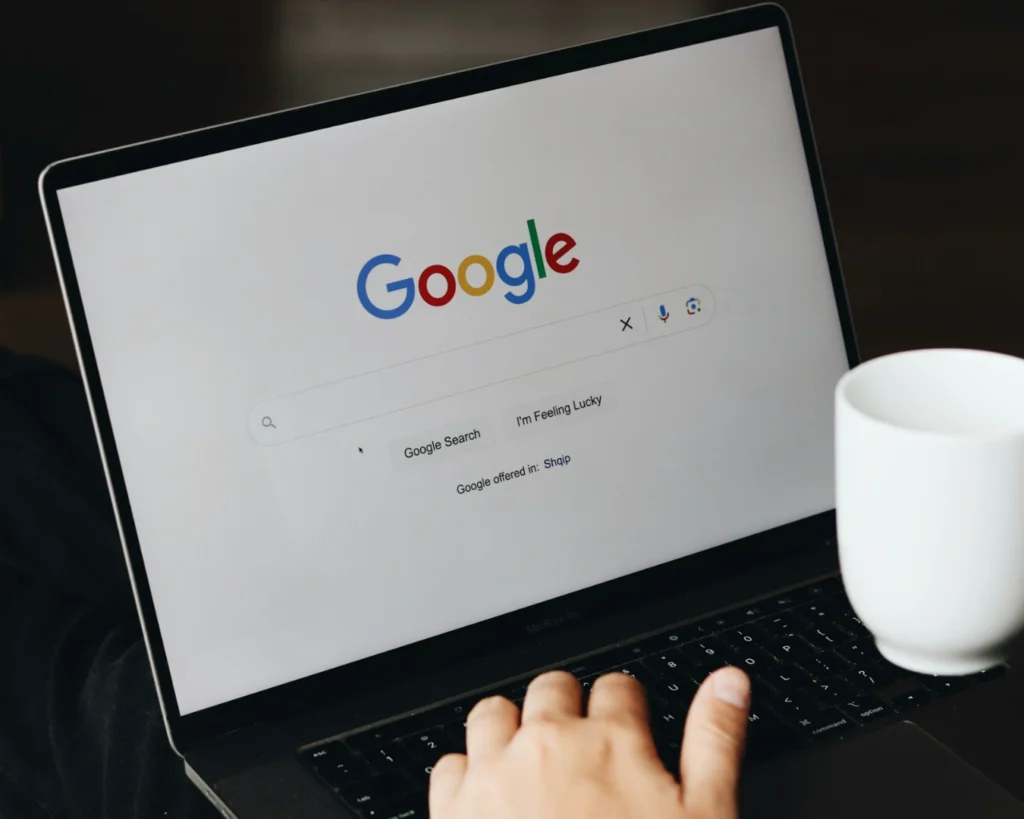 Person using a laptop displaying Google's homepage, holding a white mug, suggesting a relaxed browsing experience. Keywords: Google, laptop, coffee.