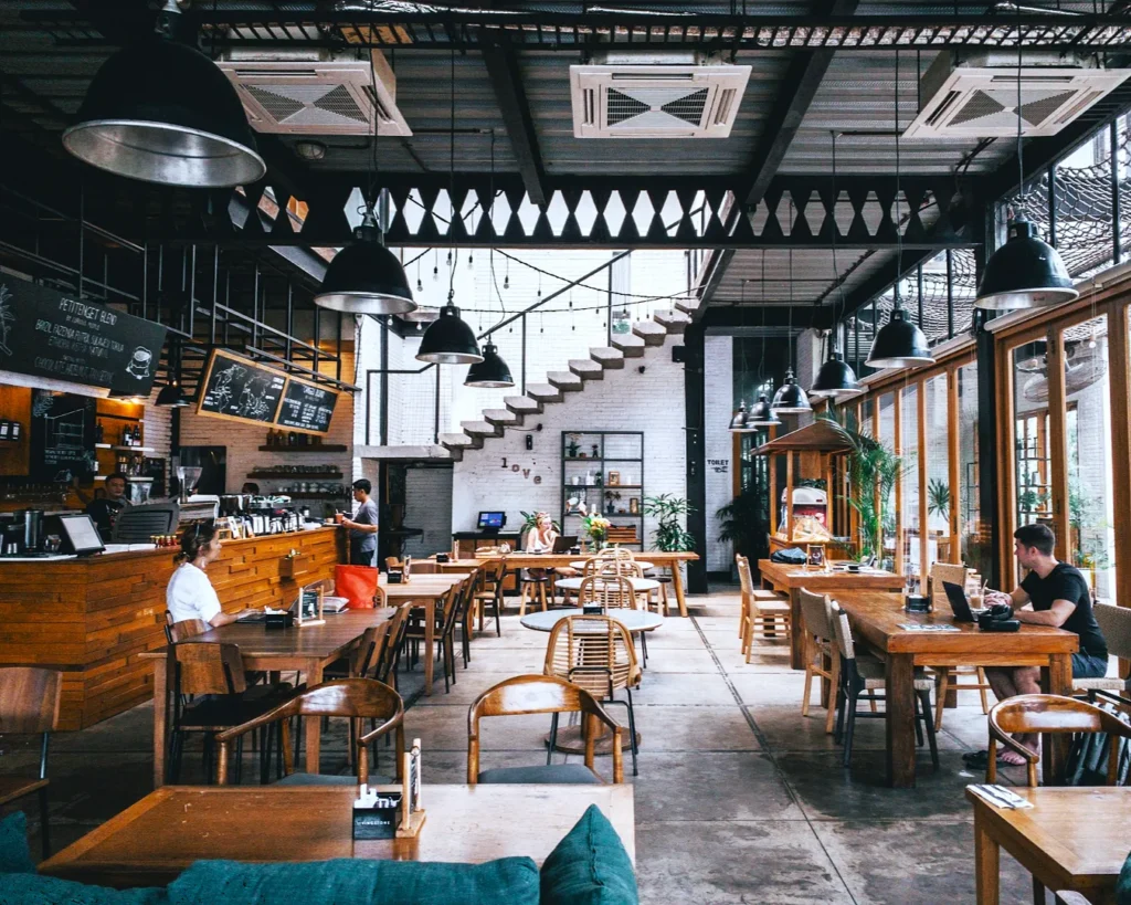 Spacious, modern cafe with wooden tables, pendant lights, and large windows. People work and socialize, creating a relaxing, inviting atmosphere.