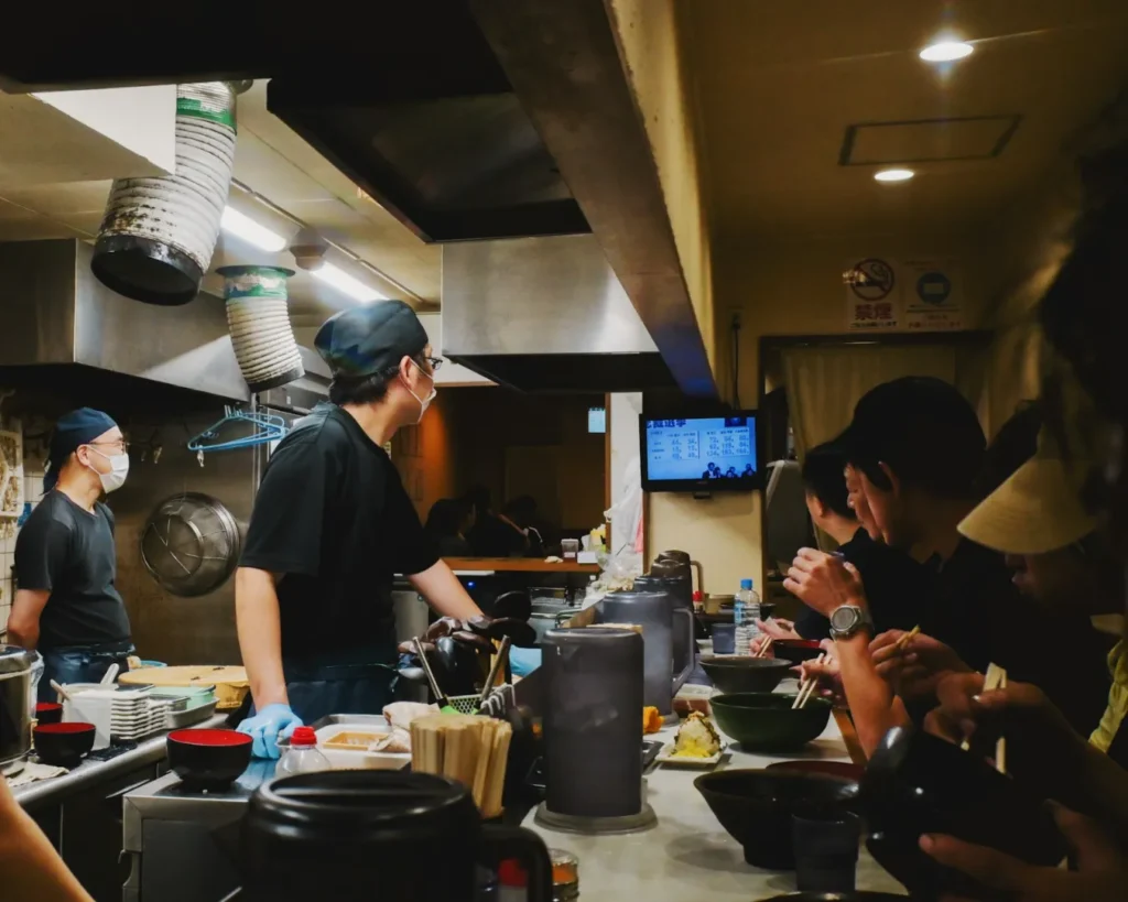A bustling ramen shop kitchen with chefs wearing black uniforms busy at work. Diners enjoy bowls of ramen at a counter, creating a lively, cozy atmosphere.