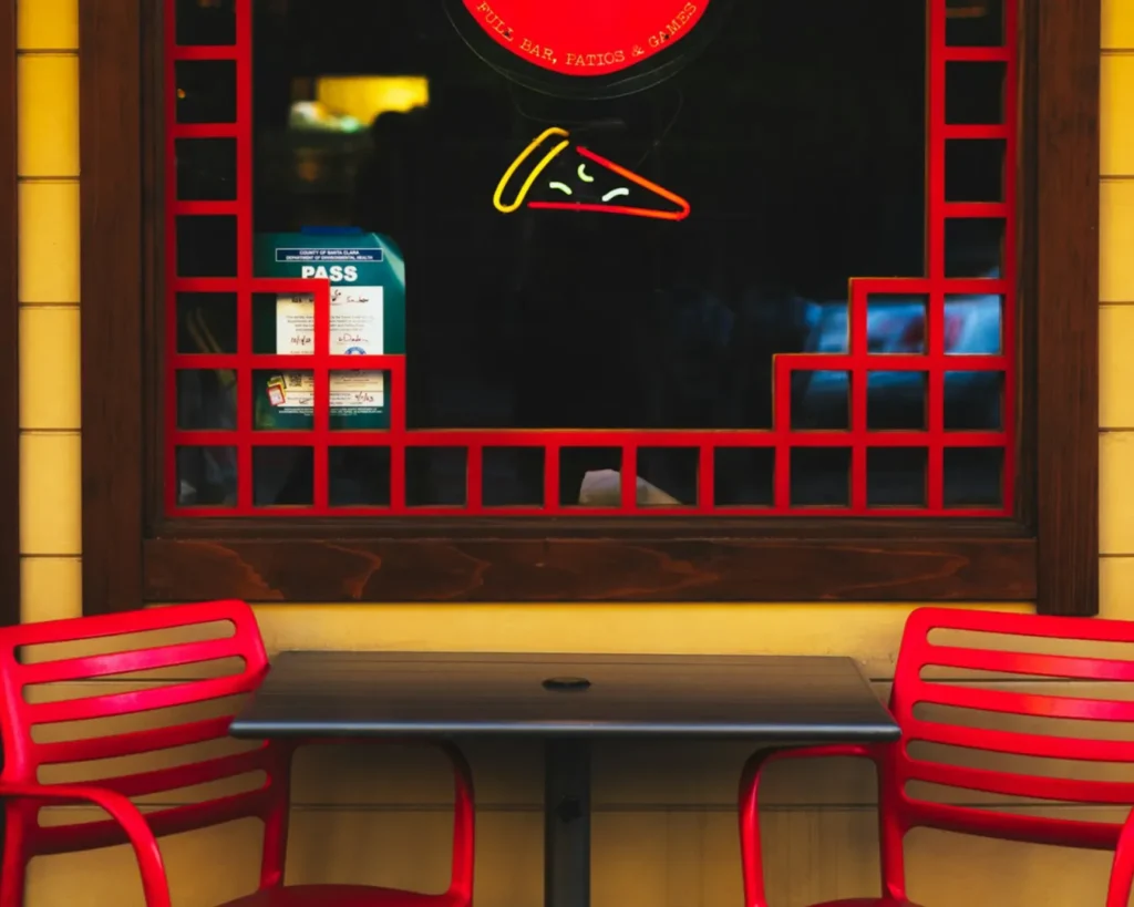 Outdoor bistro set with two red chairs and a small table in front of a window. A neon pizza slice glows inside, creating a cozy, inviting vibe.