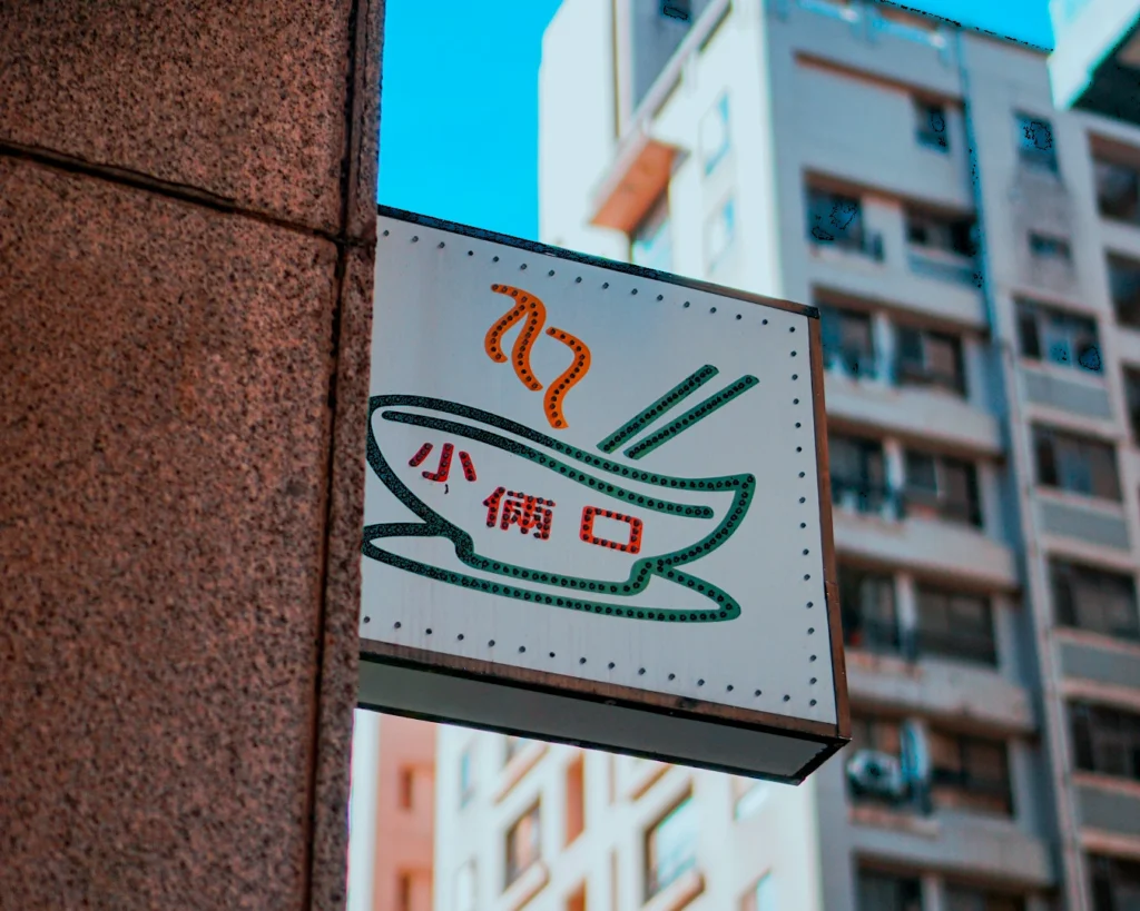 A restaurant sign features a steaming bowl of noodles and chopsticks, with text in Chinese. The sign is set against a backdrop of urban high-rise buildings.