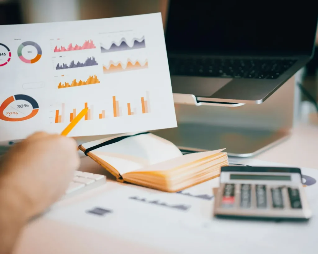 A person holds a paper with colorful charts and graphs at a desk with a laptop, notebook, and calculator, conveying a busy and analytical tone.