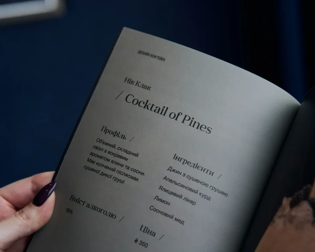 Close-up of a menu page titled "Cocktail of Pines" in elegant black font. Contains cocktail profile, ingredients, and price. Person's hand holding the page.