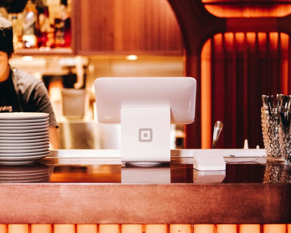 A sleek cafe counter features a glossy white POS stand with a tablet screen, a tall stack of white plates to the left, and a crystal utensil jar on the right, all bathed in warm lighting.
