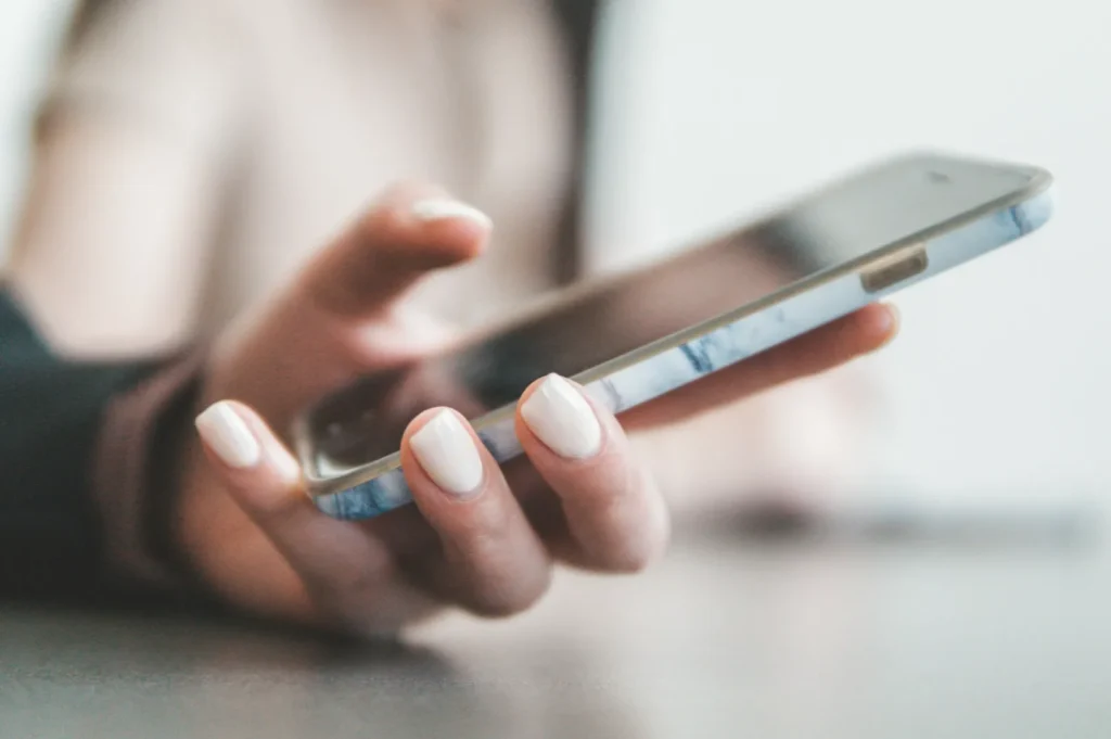 A person with manicured nails holds a smartphone, focusing on the device in a casual, relaxed setting. The image conveys a modern, connected lifestyle.