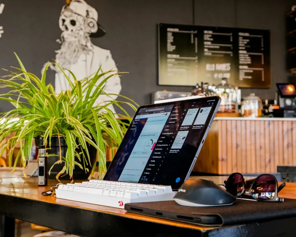 A laptop displaying a website sits on a cafe table with a keyboard and mouse. Sunglasses and a plant are nearby. A mural and menu are in the background.