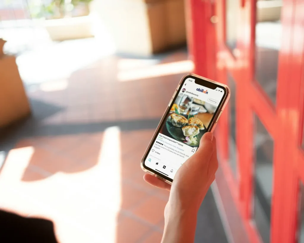 A person holds a smartphone displaying a food app with a photo of delicious empanadas. Sunlight casts soft shadows on red and brown tiled flooring.