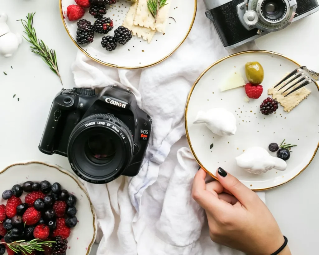 A flat lay of a Canon camera and a vintage camera with plates of berries, crackers, and herbs on a white cloth. A hand holds a plate, creating a cozy, creative vibe.