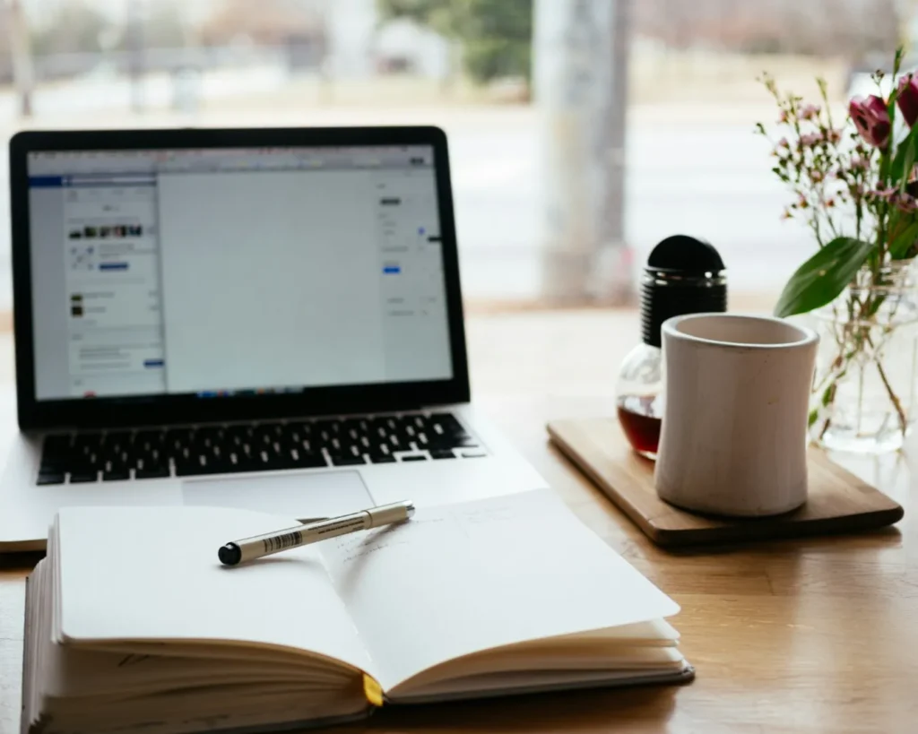 Laptop on a wooden table with an open notebook and pen in the foreground. A mug, small pot, and flowers beside them. Bright, cozy workspace.