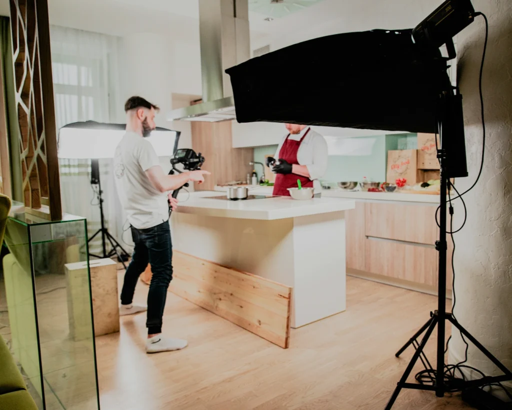A photographer films a cooking show in a bright kitchen. Lights and camera equipment surround a chef preparing food on an island counter.