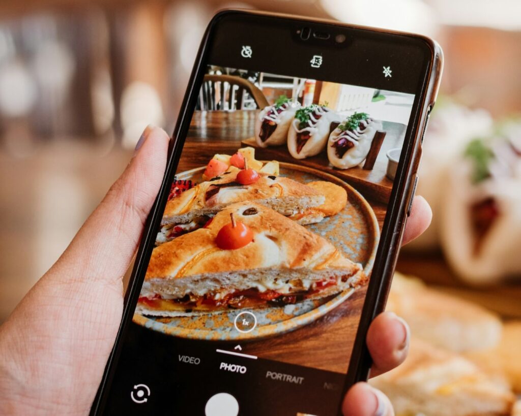 A hand holds a smartphone, capturing a photo of gourmet sandwiches filled with vegetables and wraps topped with greens. The scene conveys a warm, appetizing ambiance.