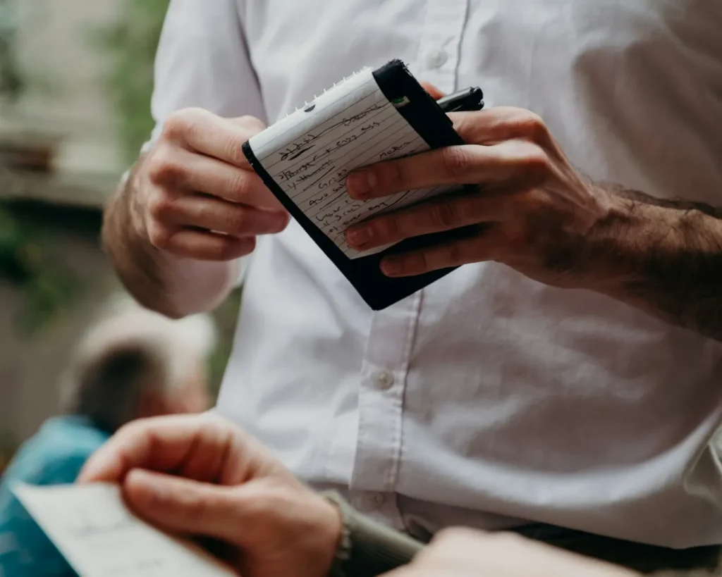 A person in a white shirt is holding a notepad and pen, seemingly taking an order from another person. The scene appears calm and professional.