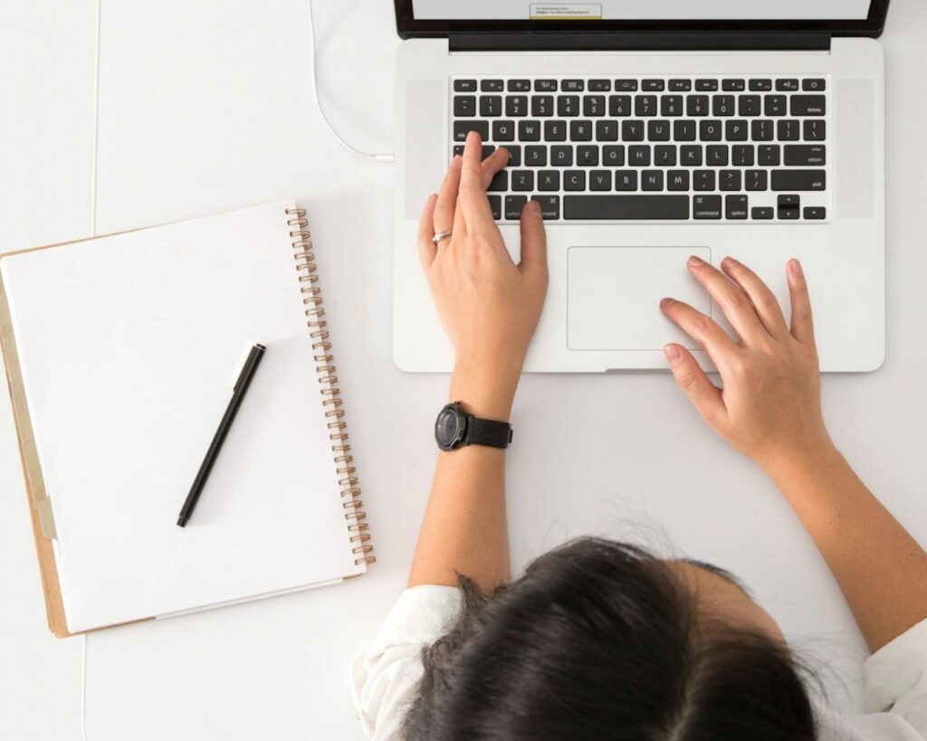 Aerial view of hands typing on a laptop, with an open notebook and pen nearby on a white workspace.