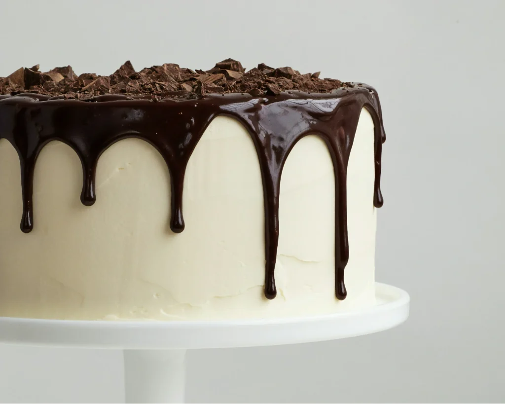 A cake with white frosting, topped with chocolate shavings and dripping dark chocolate glaze, sits elegantly on a white pedestal stand.
