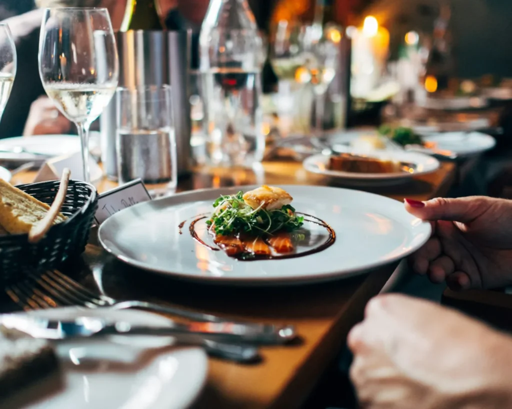 Elegant dining scene with a hands holding a white plate featuring gourmet food garnished with greens and sauce. Glasses of white wine and bread in a basket add to the sophisticated ambiance.