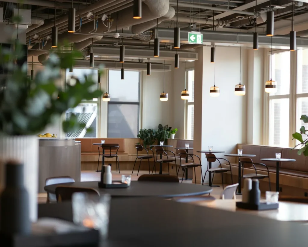 Modern cafe interior with wooden tables, black chairs, and green plants by large windows. Minimalistic lighting creates a calm, inviting atmosphere