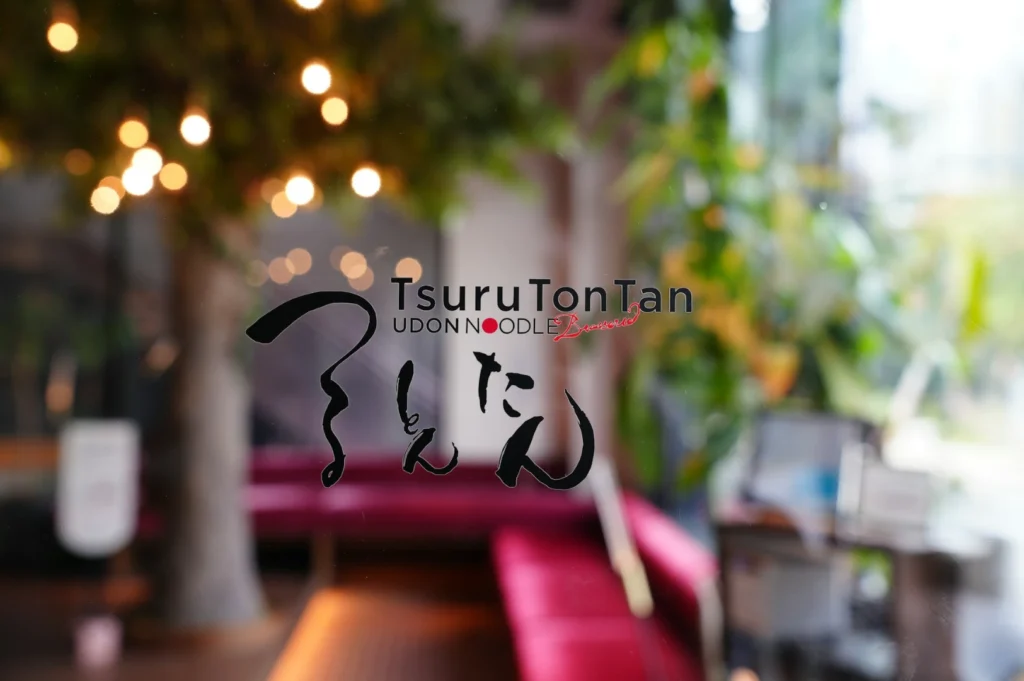Blurred view of a cozy restaurant interior with warm lighting and green plants. "TsuruTonTan Udon Noodle Brasserie" is written on the glass in foreground.