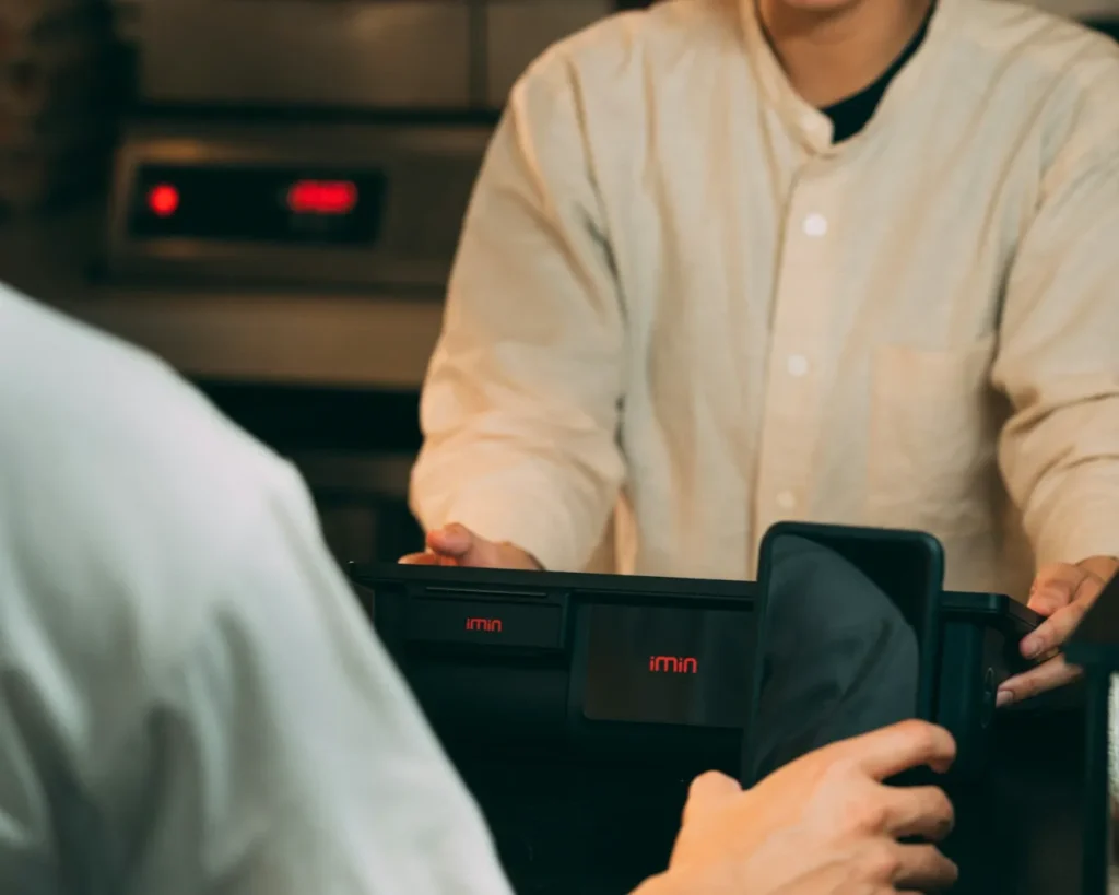 A person in a beige shirt stands at a counter, holding a black device, as another hand points a smartphone towards it, suggesting a digital payment.